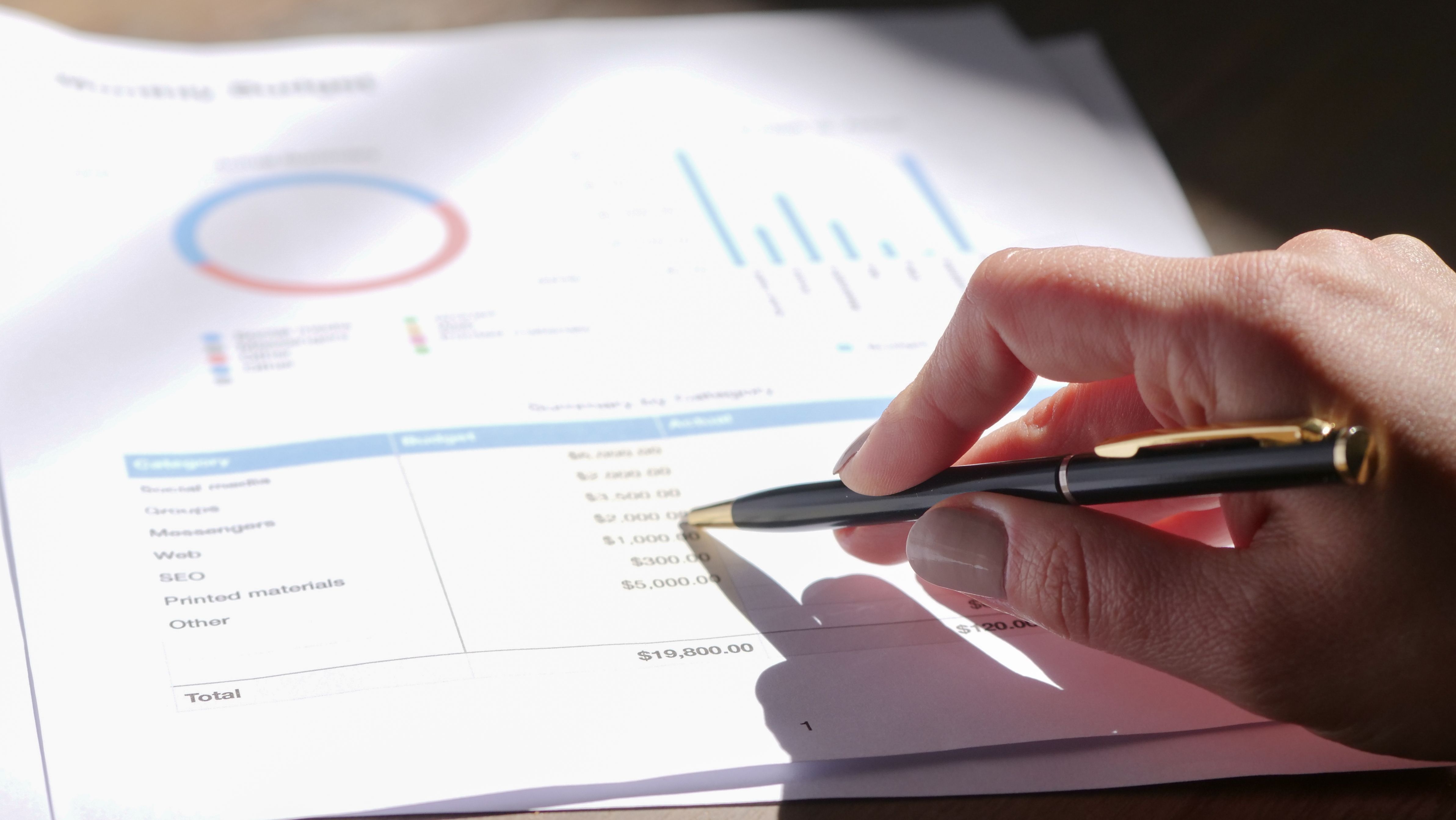 Hand holding a pen while analyzing a budget with charts on graph paper